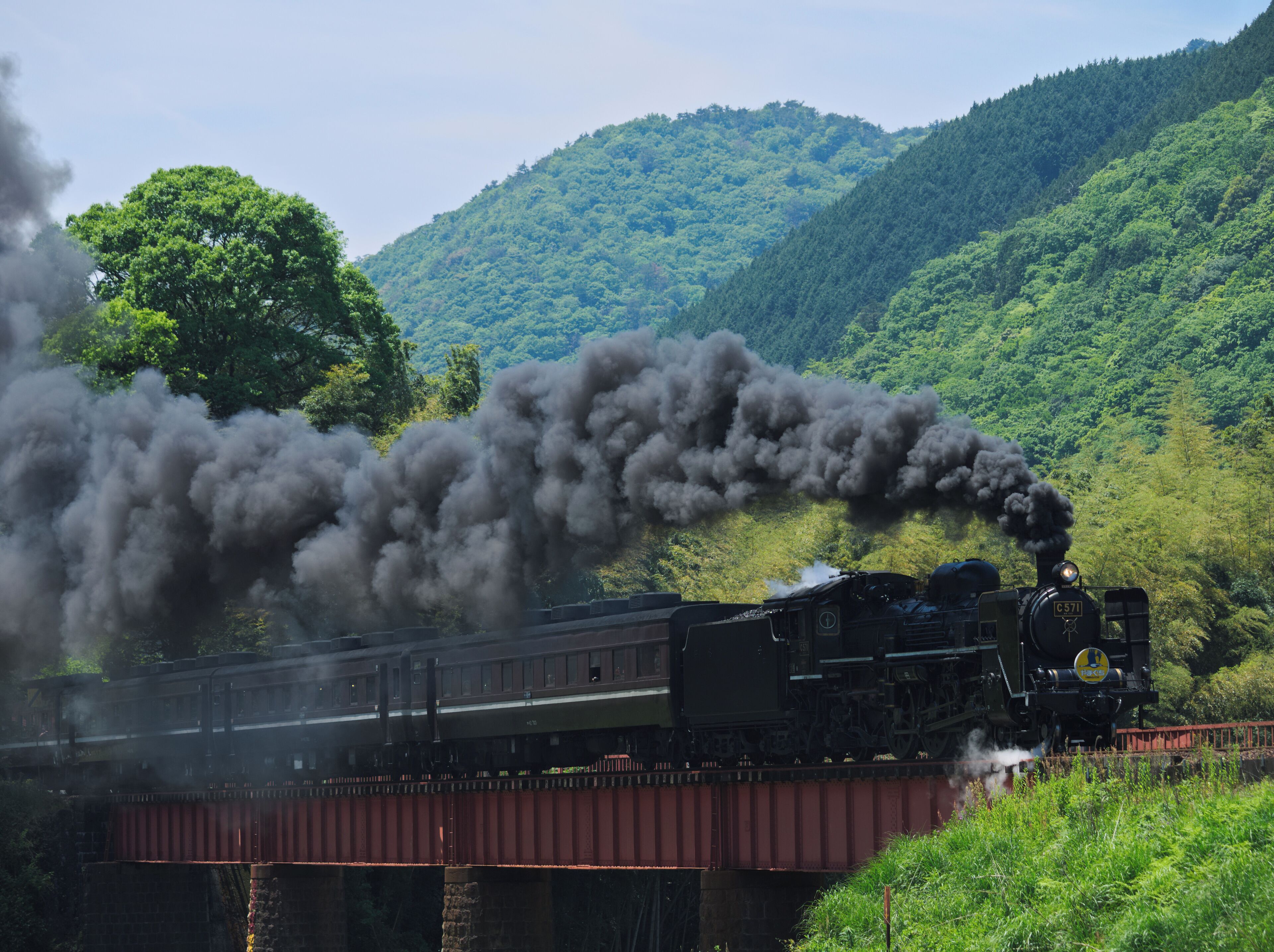 A steam locomotive emitting thick black smoke, crossing a bridge over a lush green valley with dense forested hills in the background.