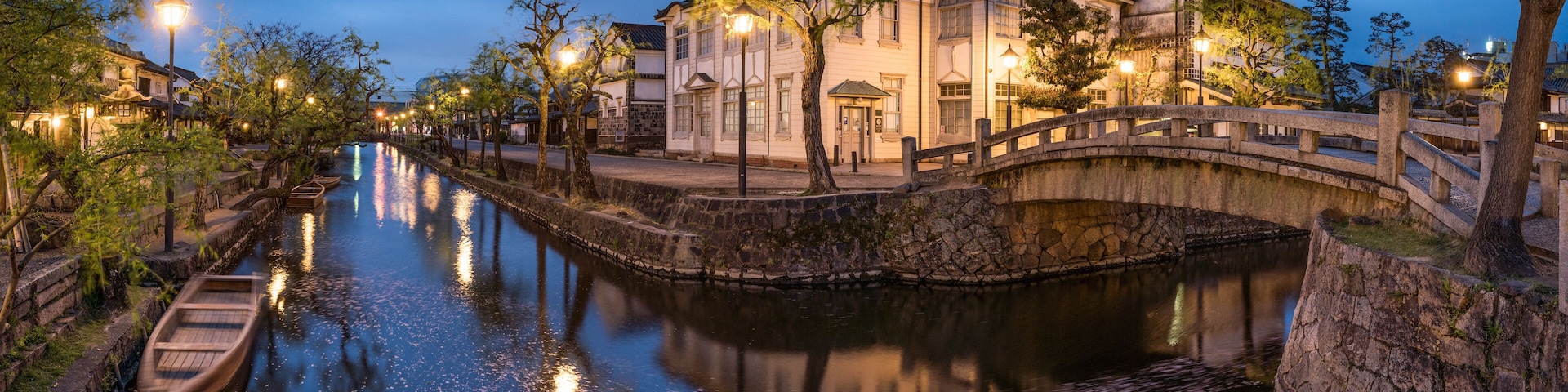Um canal com um barco de madeira, uma ponte de pedra e edifícios tradicionais ao entardecer.