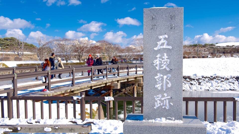 A stone monument with Chinese characters, a wooden bridge over a river, and a snowy landscape with people walking.