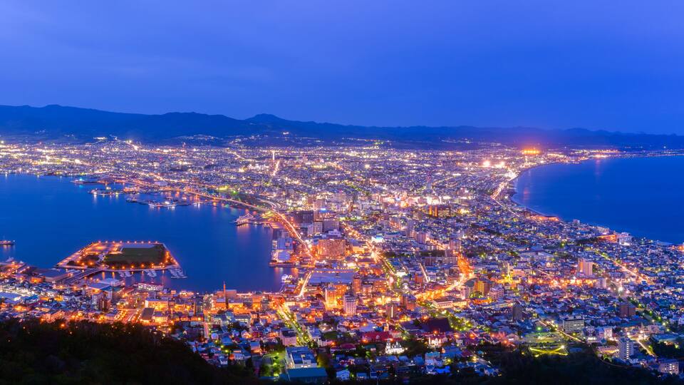 A coastal cityscape at dusk with illuminated buildings, a marina, and a large body of water.