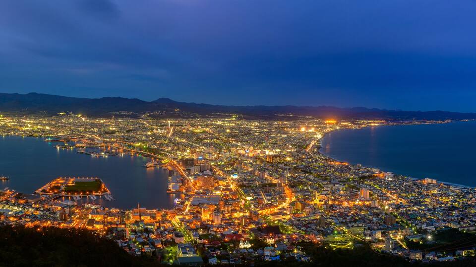 A coastal cityscape at night with illuminated buildings, a marina, and mountains in the background.