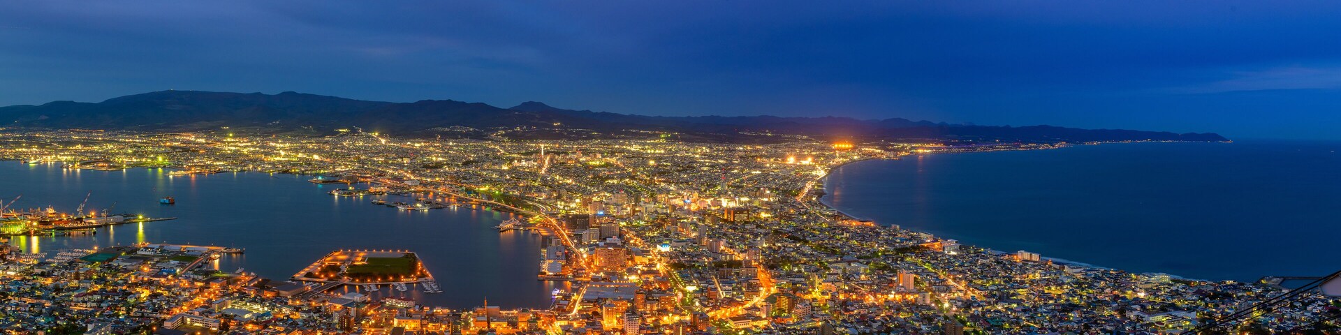 Un paisaje urbano costero nocturno con edificios iluminados, un puerto deportivo y montañas al fondo.
