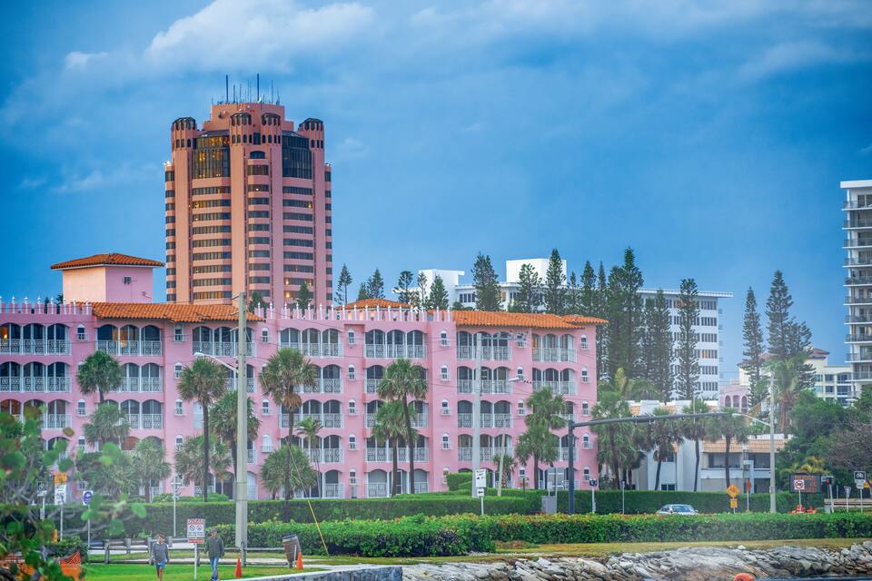A pink building with a red roof, surrounded by palm trees and other buildings.