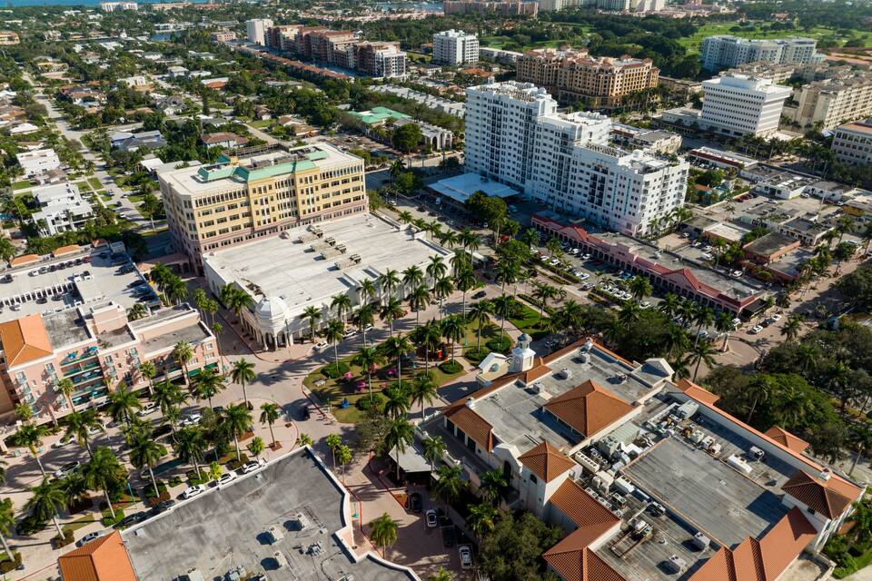 An aerial view of a city with dense urban development, including high-rise buildings, a large parking area, and palm trees.