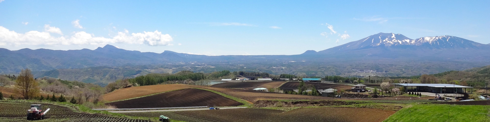 A vast agricultural field with rows of young plants, a tractor, and a blue vehicle. In the background, there are buildings and a snow-capped mountain.