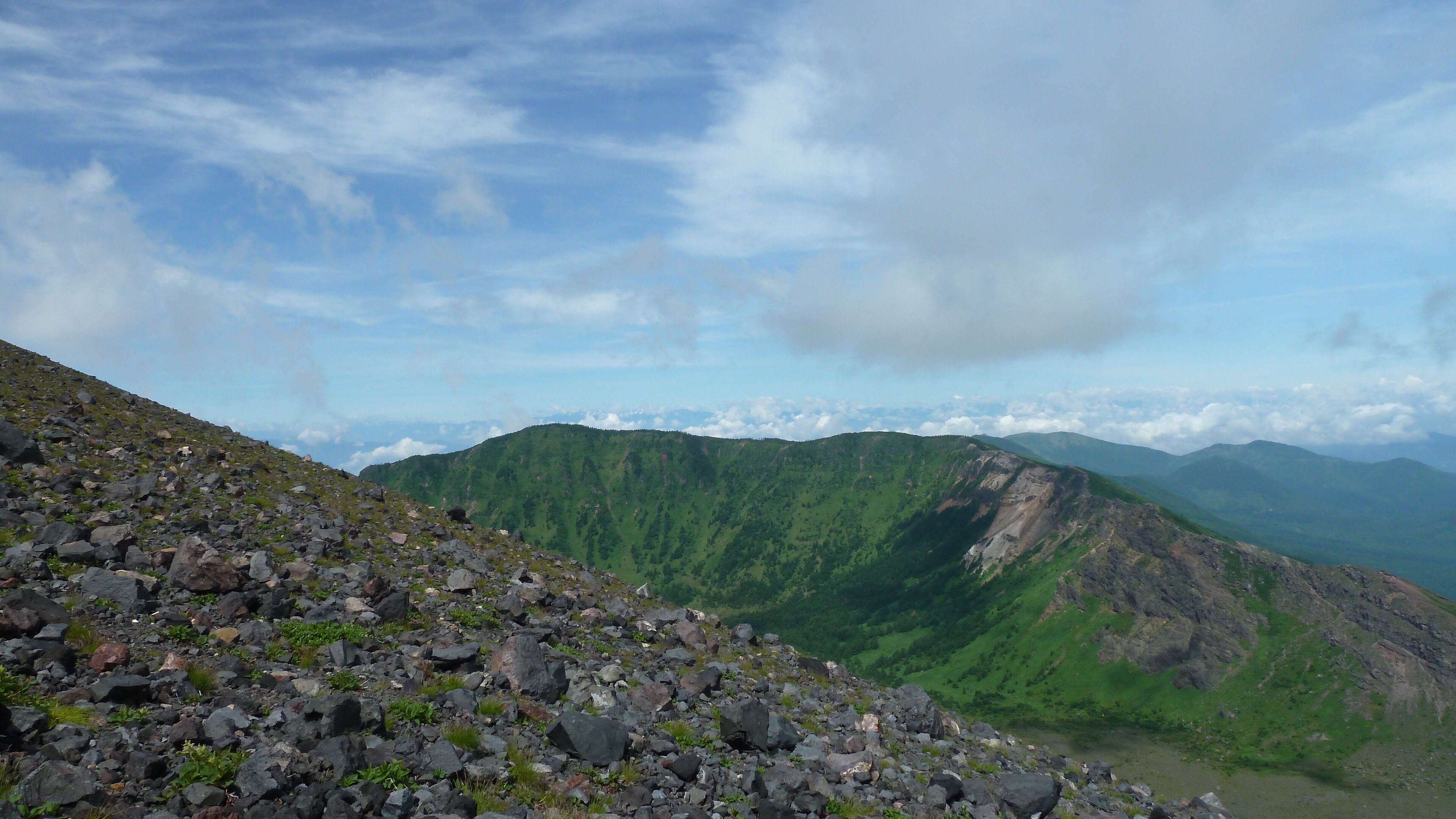 A rocky mountain slope with a valley and distant mountains under a partly cloudy sky.