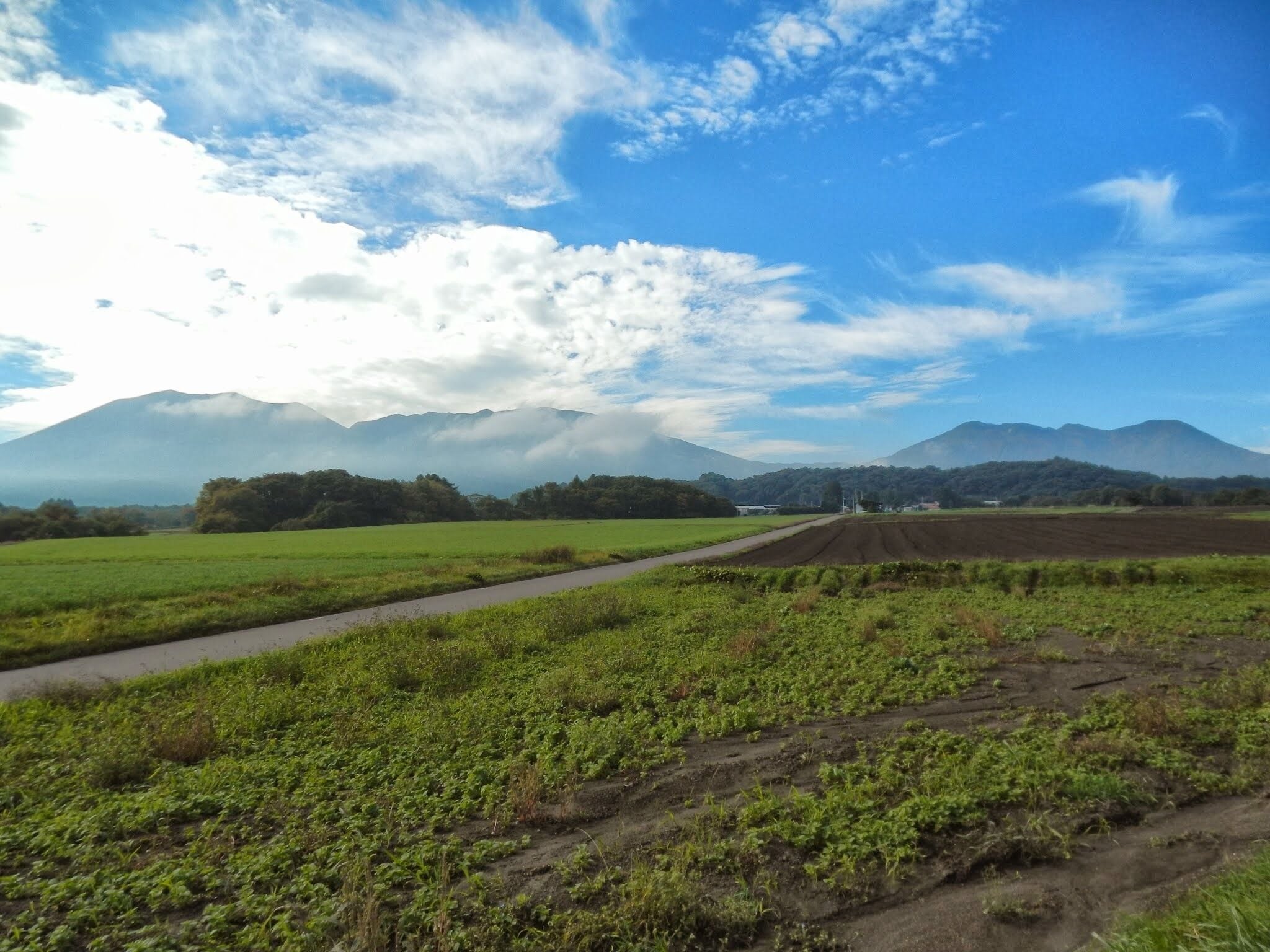 A rural landscape with a dirt path, green fields, and distant mountains under a partly cloudy sky.