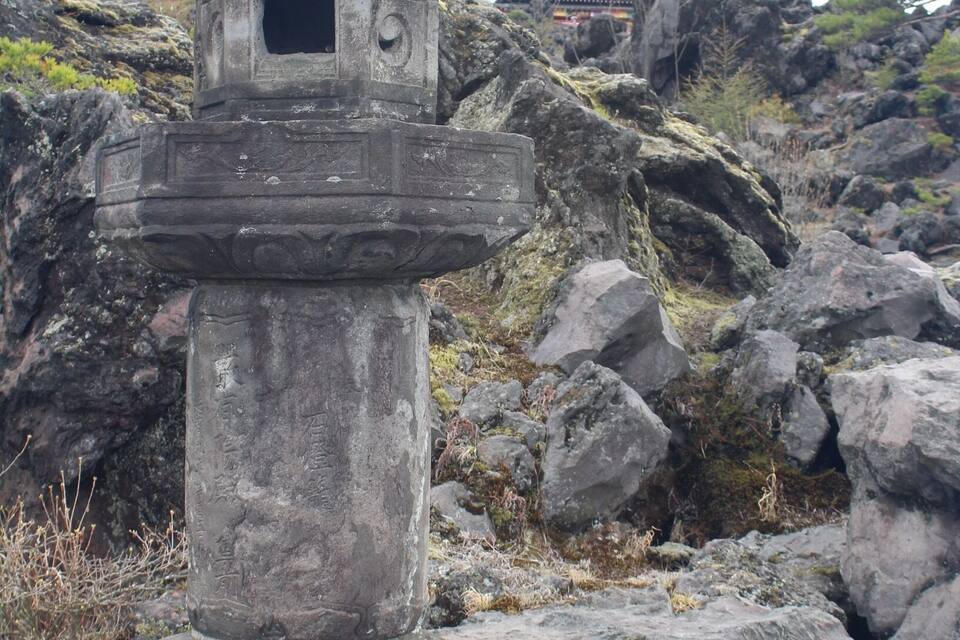 A stone lantern with a traditional design, set against a rocky landscape with a building in the background.