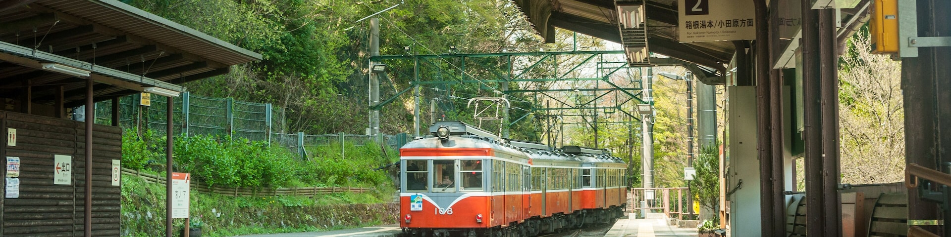 Um trem em uma estação com plataforma coberta, bancos e vegetação ao fundo.