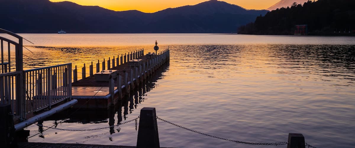 A pier extending into calm waters with a sunset and mountains in the background.