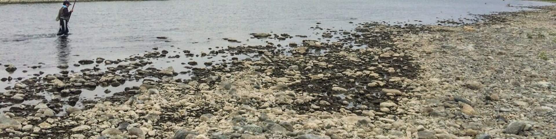 A person fishing by a rocky riverbank with a calm body of water and a cloudy sky.