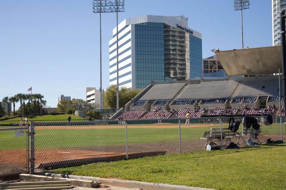 Un campo da baseball con gradinate vuote, una rete metallica e un edificio moderno sullo sfondo.
