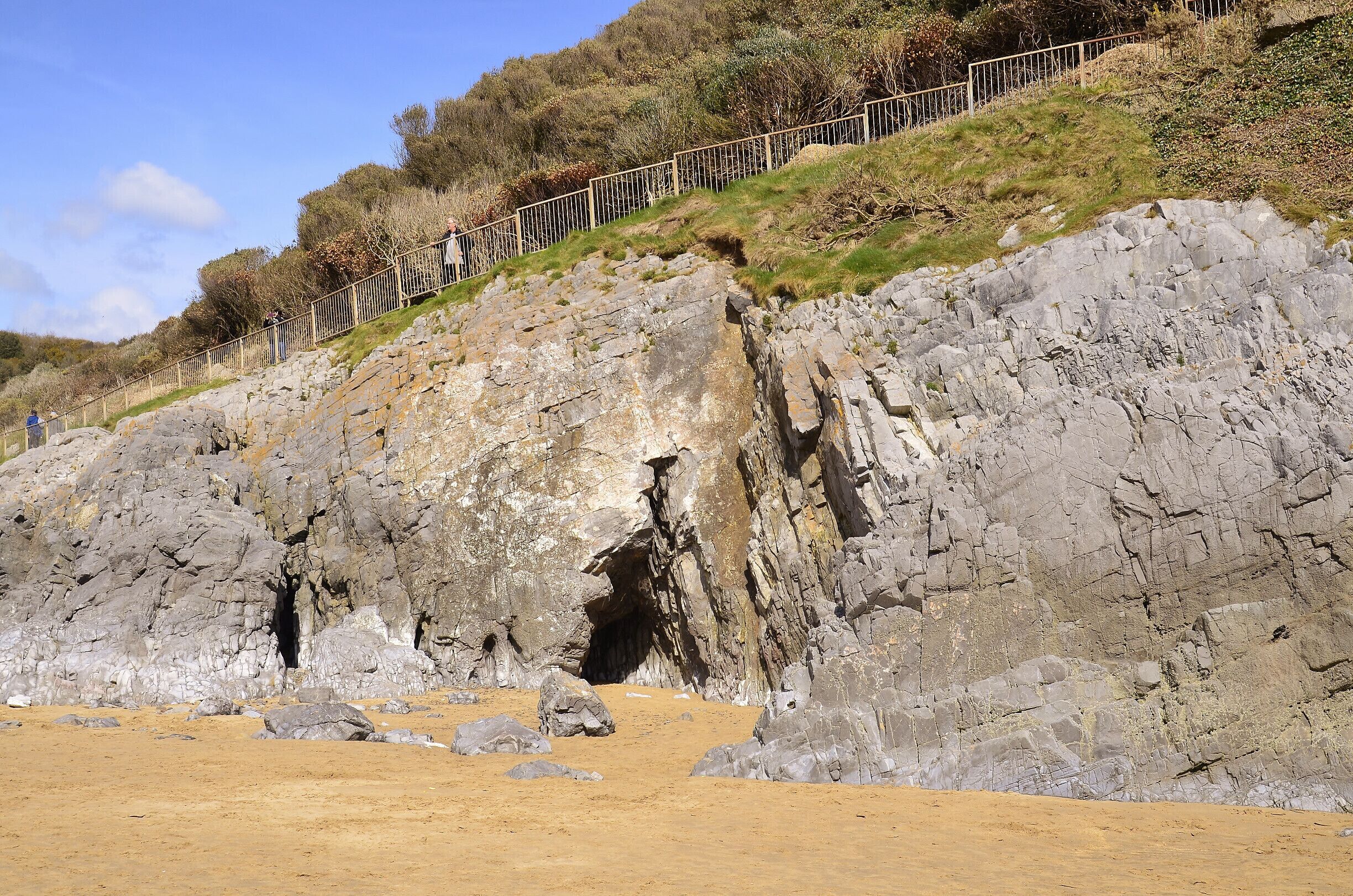A rocky cliff with a cave entrance, a sandy beach, and a metal fence along the top.