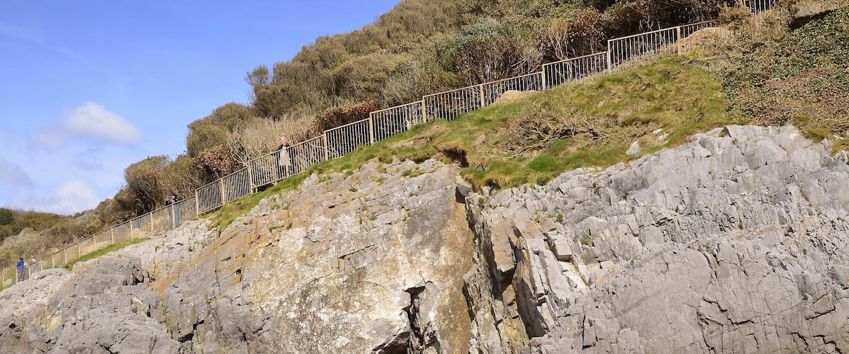 A rocky cliff with a cave entrance, a sandy beach, and a metal fence along the top.