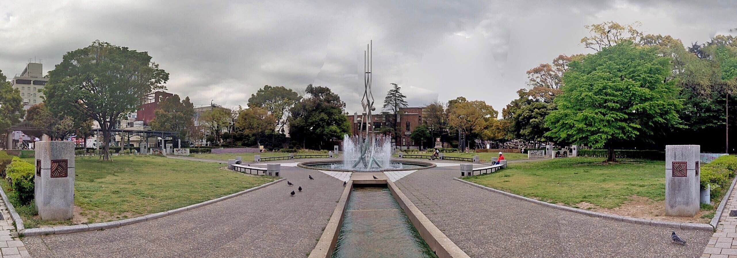 Um parque com uma fonte central, rodeado por árvores e um passeio pavimentado.