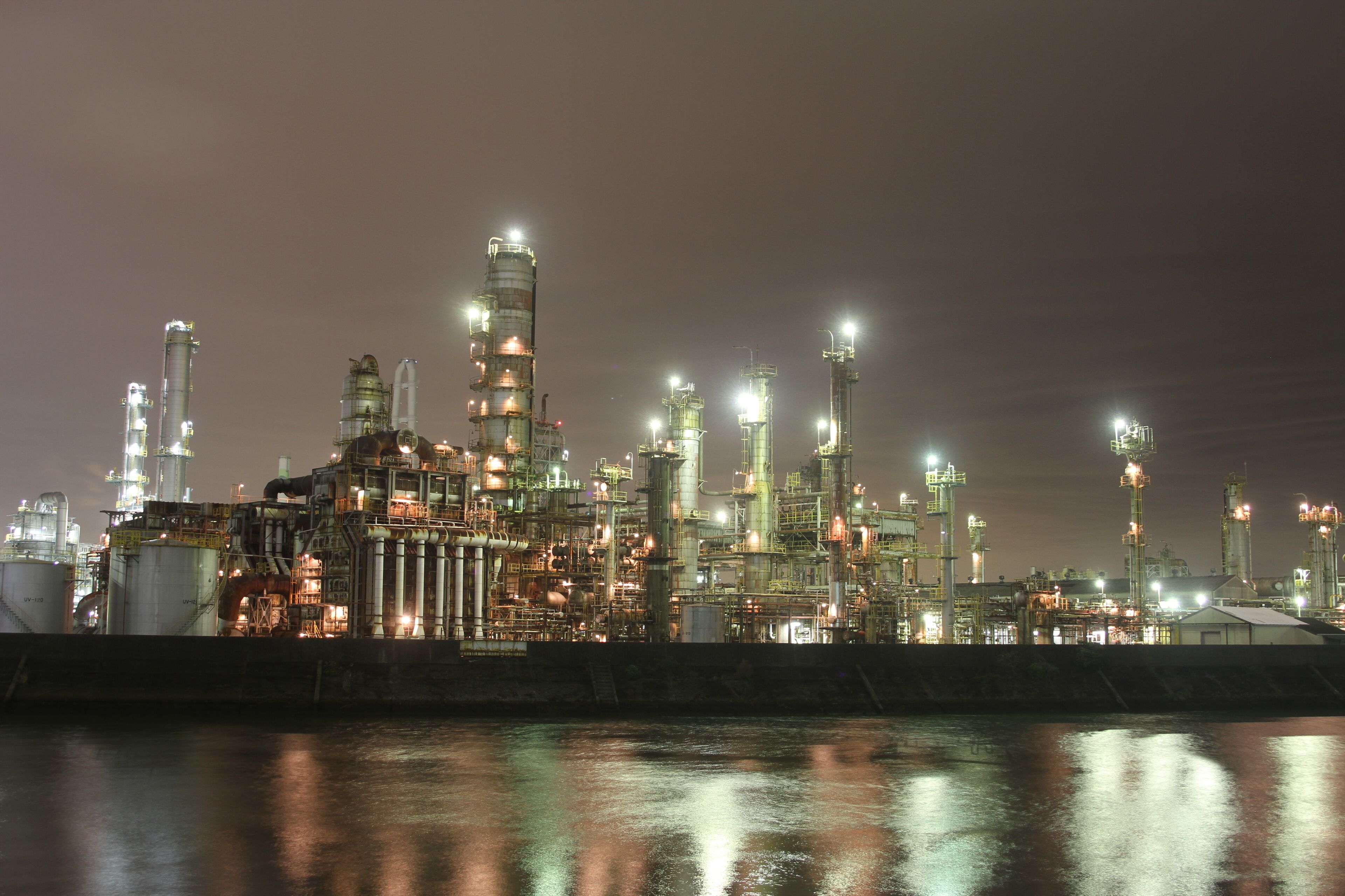 An industrial complex at night with illuminated structures and a body of water reflecting the lights.