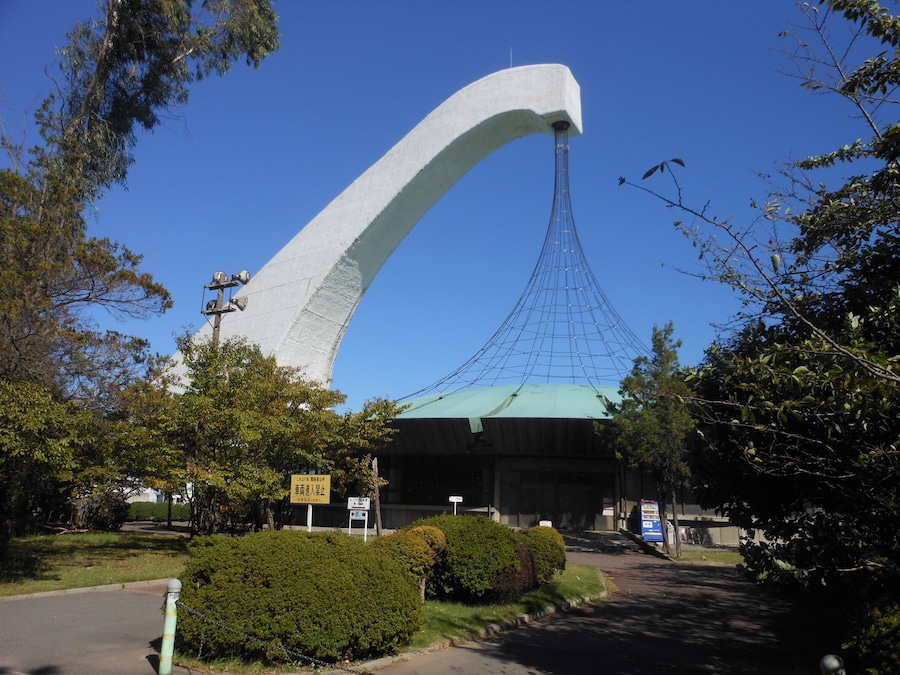 A large, curved white structure with a green roof, surrounded by trees and bushes.