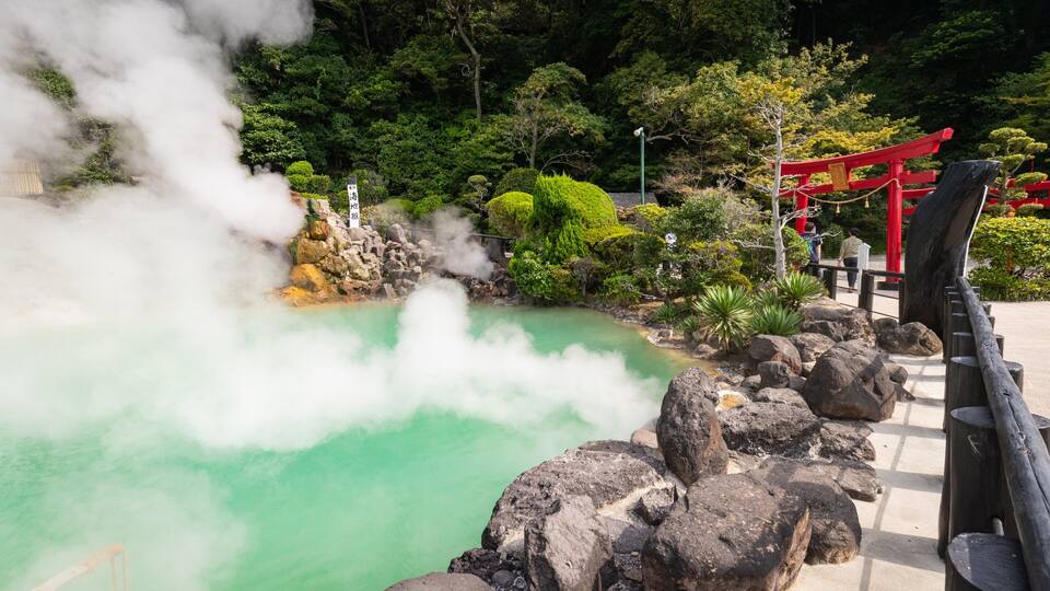 A hot spring with steam rising, surrounded by rocks and greenery, and a red torii gate in the background.