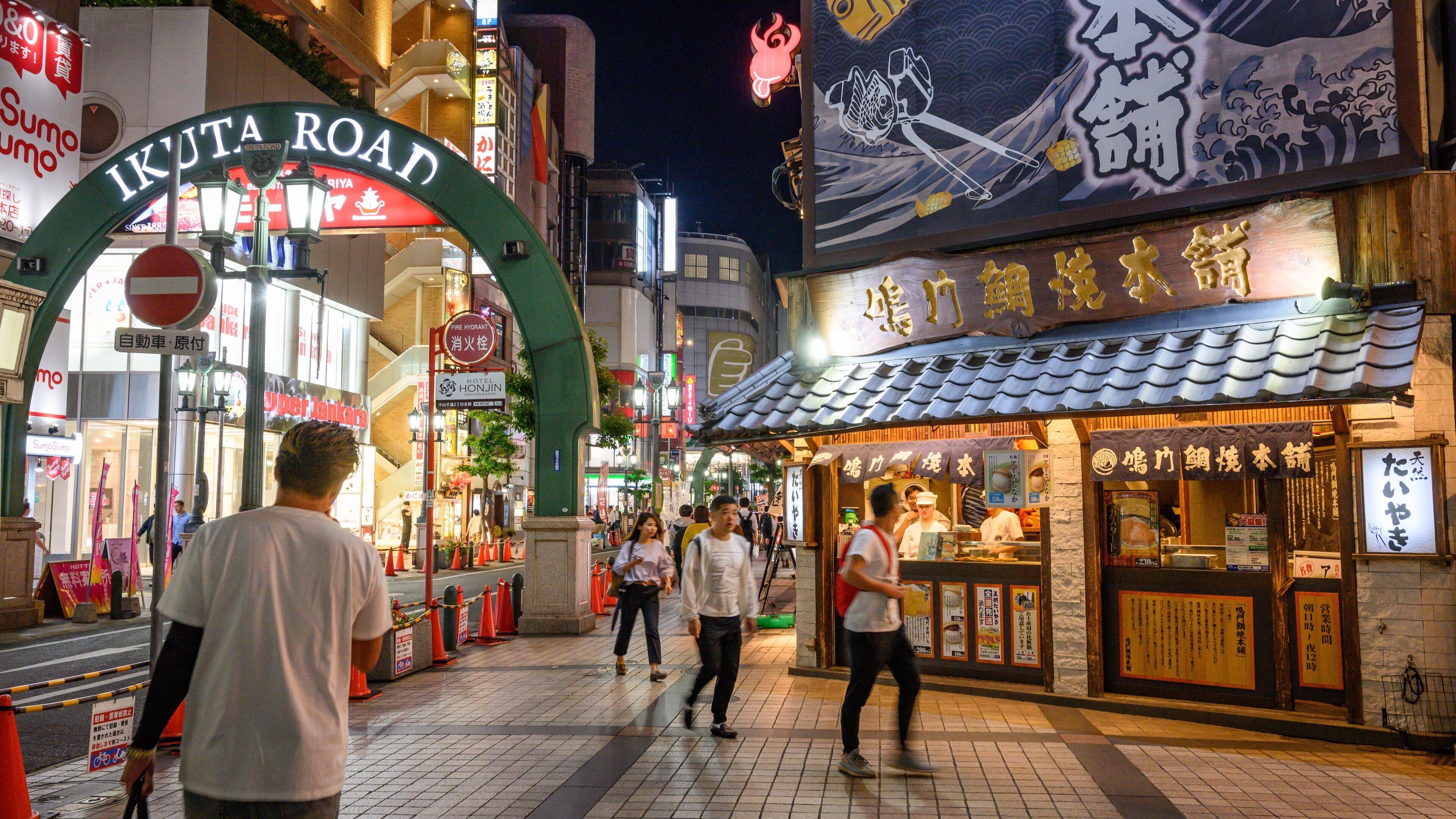 A busy street with a prominent sign for Ikuta Road, various shops, and a traditional Japanese restaurant.