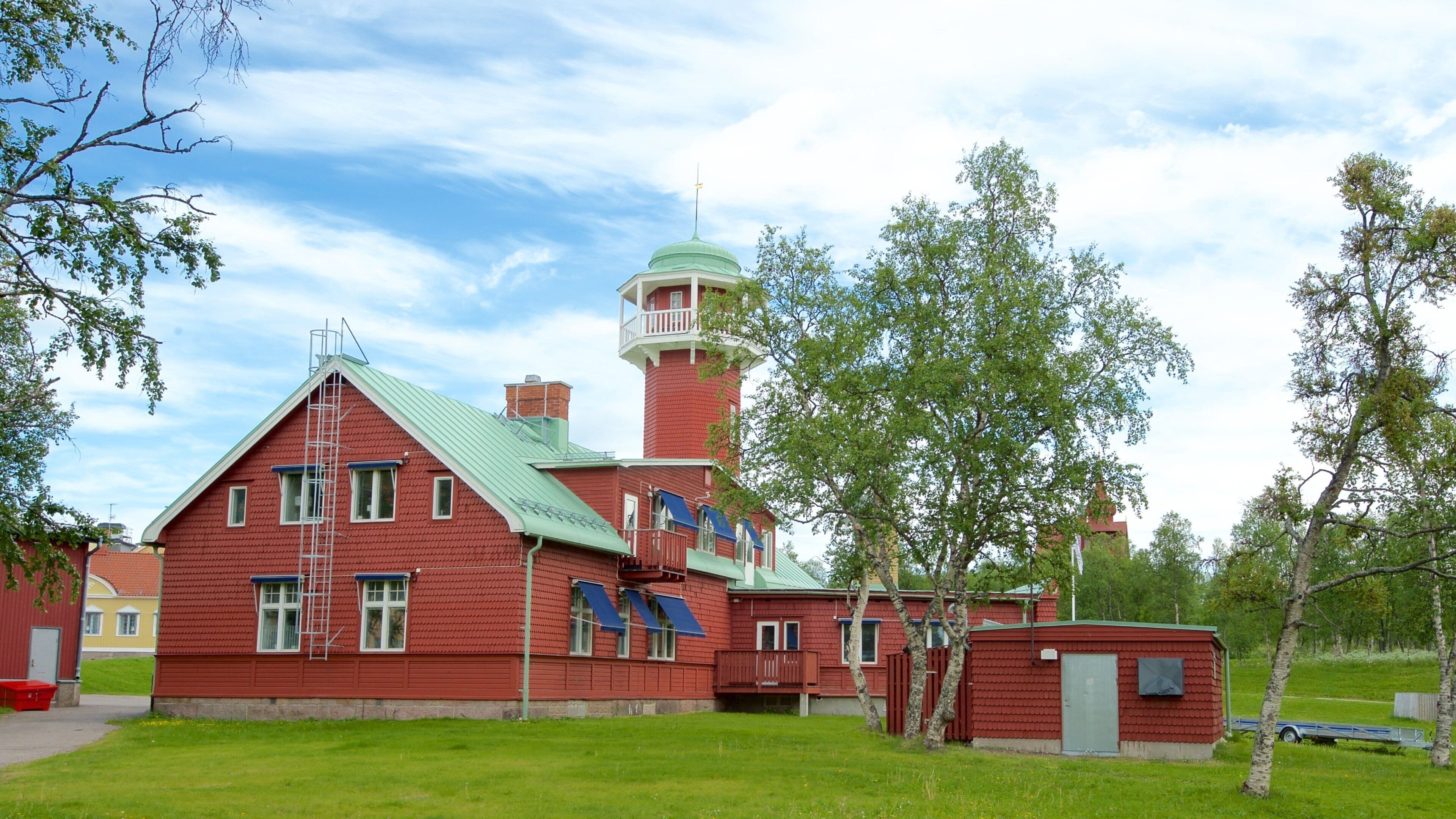 A red brick lighthouse with a green roof and a white balcony, surrounded by trees and grass.