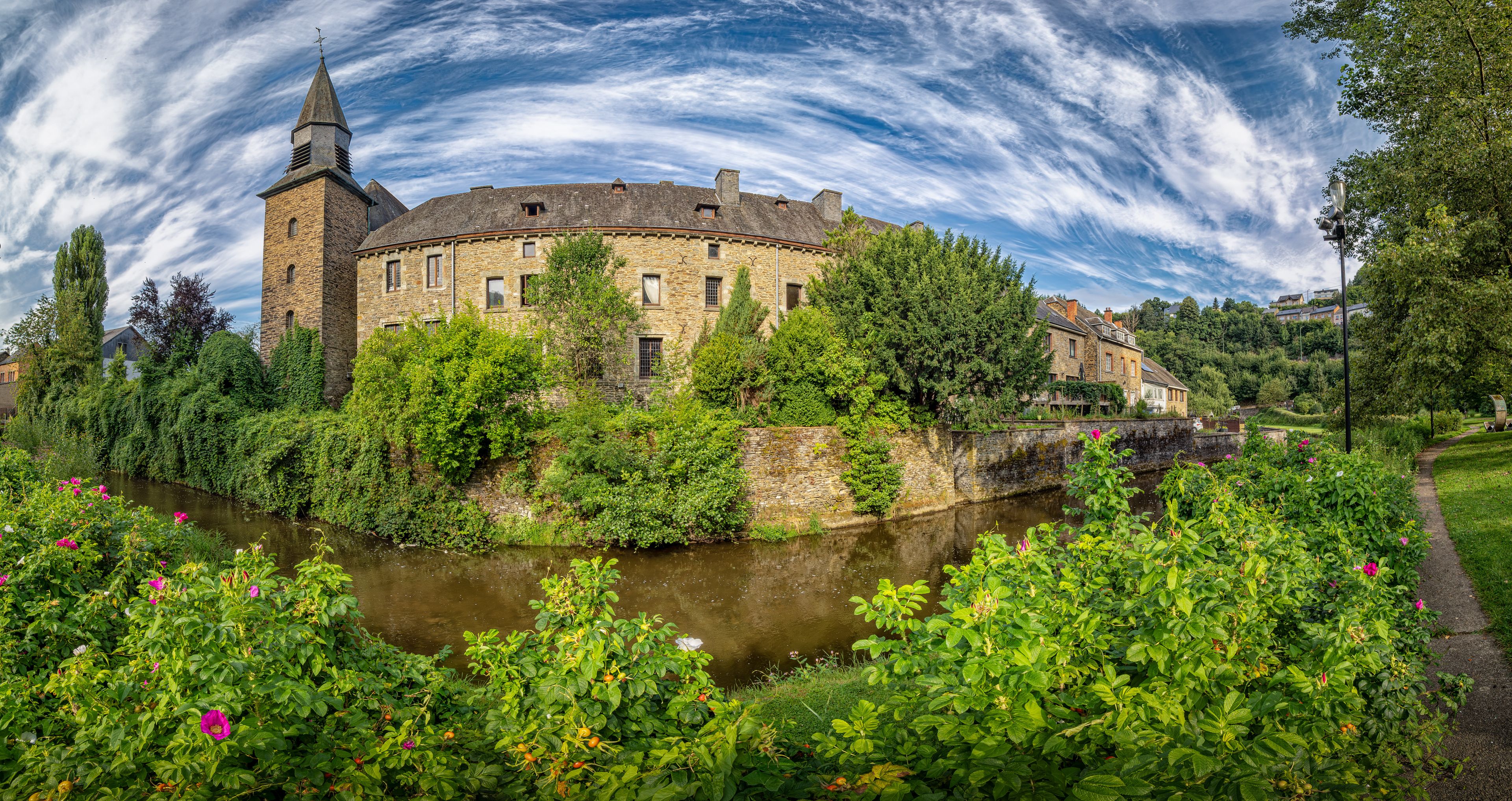 Un bâtiment historique en pierre, doté d’une tour, entouré d’une végétation luxuriante et bordé par une rivière.