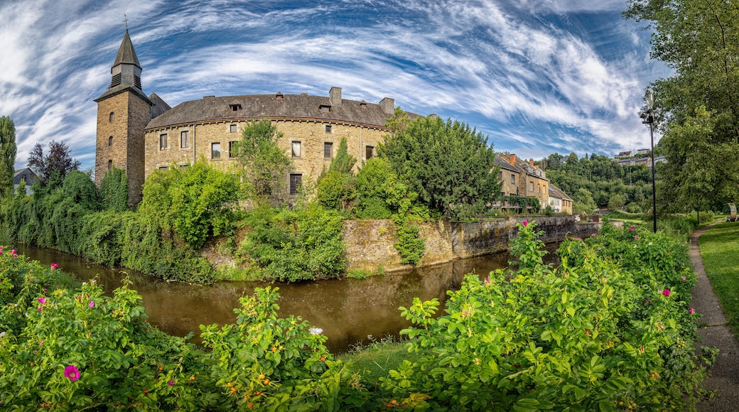 Un bâtiment historique en pierre, doté d’une tour, entouré d’une végétation luxuriante et bordé par une rivière.