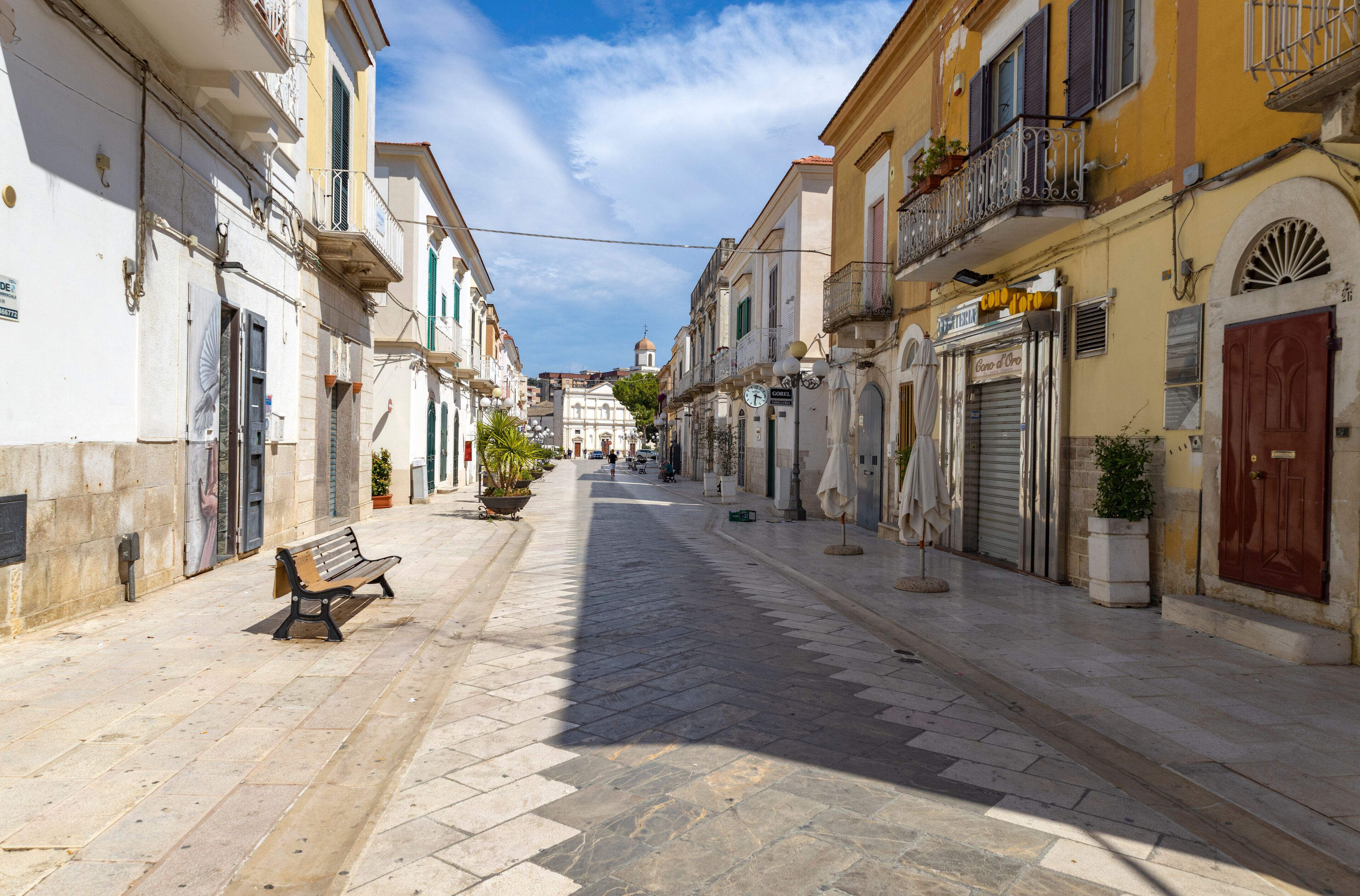 A sunlit street with a bench, buildings, and a distant dome.