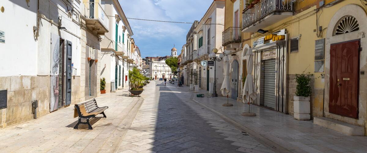 A sunlit street with a bench, buildings, and a distant dome.