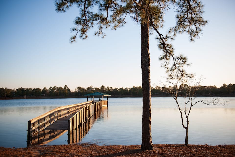 A wooden pier extending into a calm lake, surrounded by trees and a clear sky.