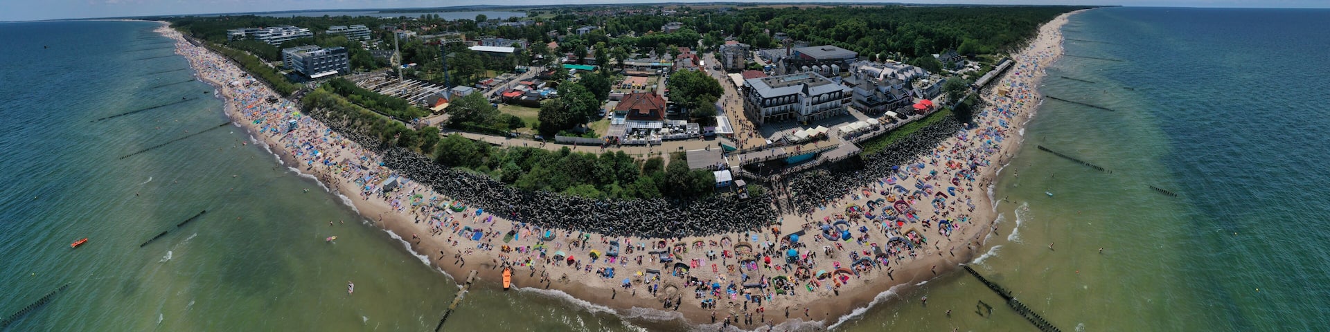 Drone aerial perspective and panoramic view on sunny beach with sunbathers with windbreaks and towels at sea in touristic city