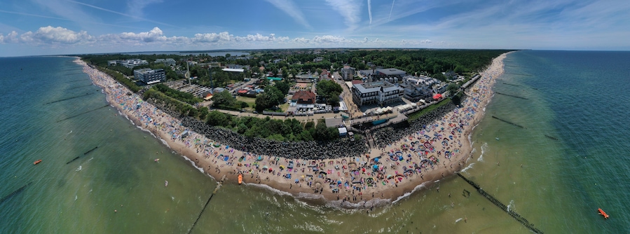 Drone aerial perspective and panoramic view on sunny beach with sunbathers with windbreaks and towels at sea in touristic city