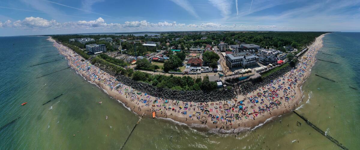 An aerial view of a beach with a dense crowd, clear waters, and buildings in the background.