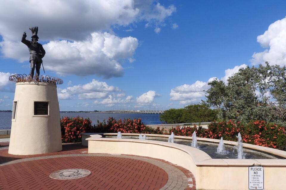 Uma estátua em um pedestal com uma placa, uma fonte e um corpo de água ao fundo.