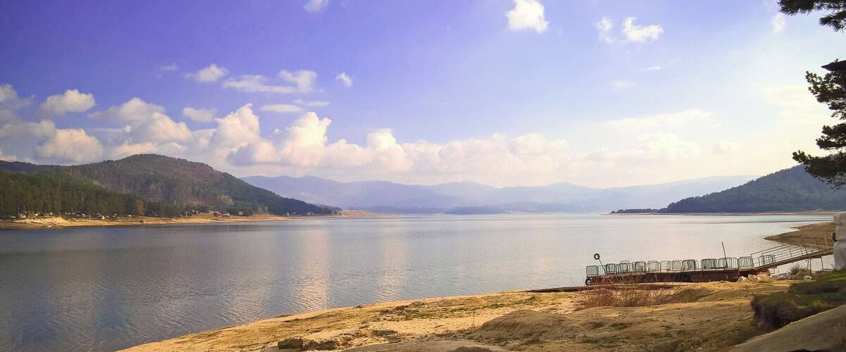 A calm lake with a sandy shore, a dock with chairs, and distant mountains under a clear sky.