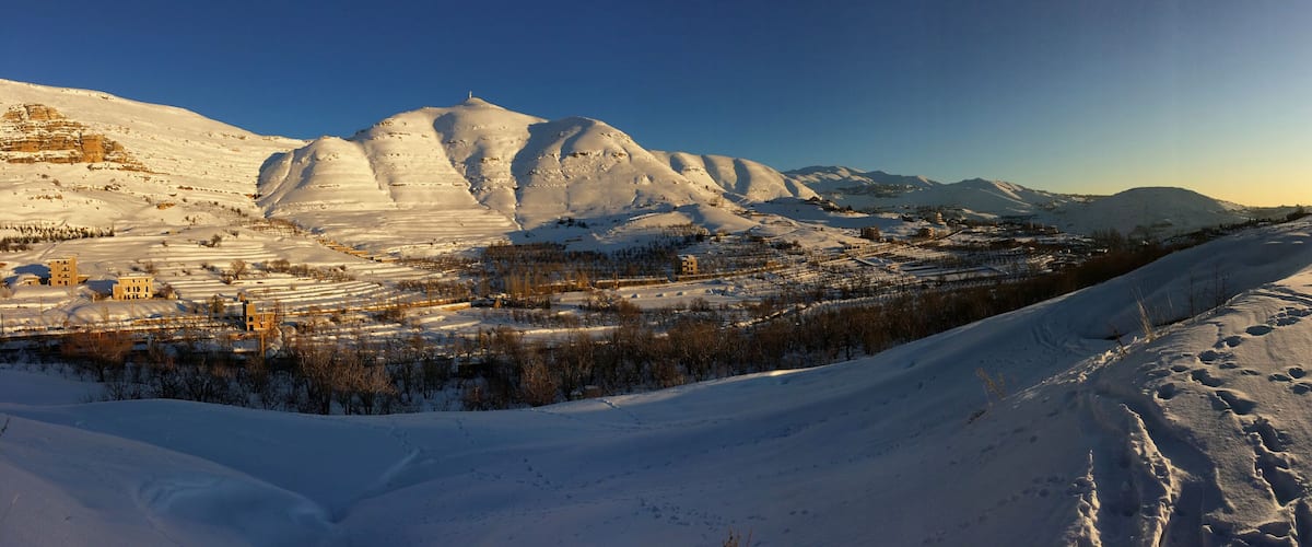 A snow-covered mountainous landscape with a clear sky and the sun setting in the background.