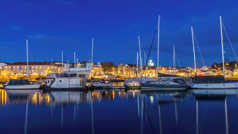 A marina at dusk with several sailboats docked, reflecting in calm waters, and buildings in the background.