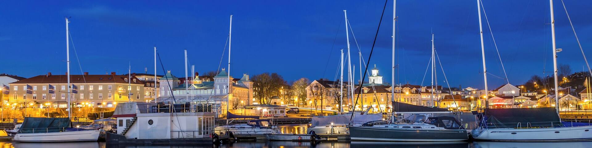 A marina at dusk with several sailboats docked, reflecting in calm waters, and buildings in the background.