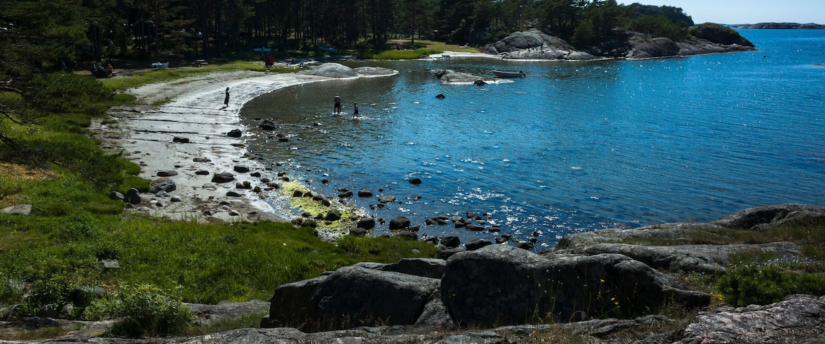 Small crescent-shaped beach on Rossö Island, Sweden. Rocky cliffs, shallow water, and a pine forest meet the blue sea under a clear summer sky. A serene coastal landscape