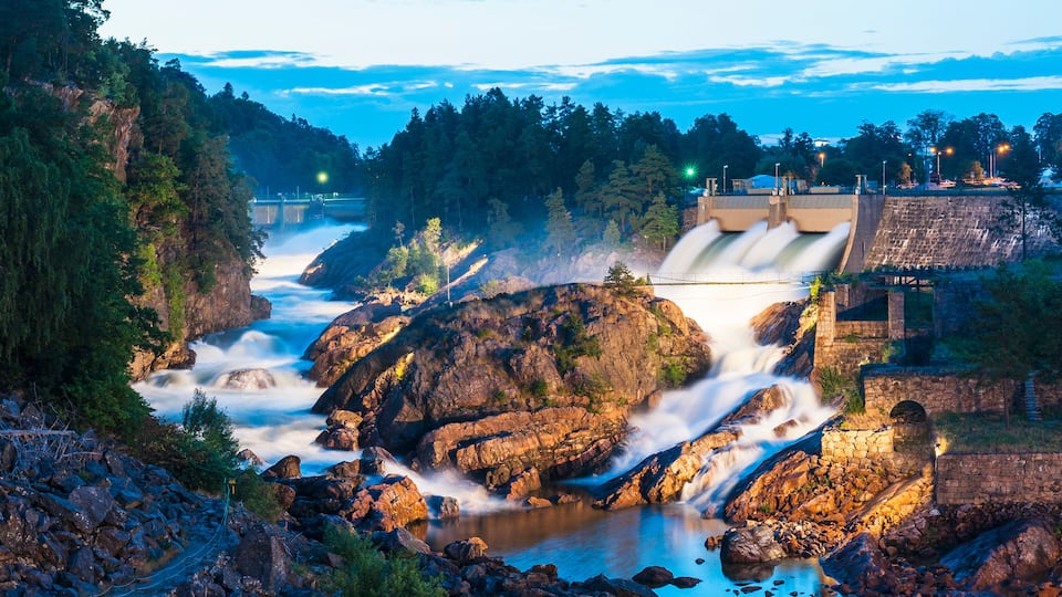 A dam with water cascading over rocks, a forested hillside, and a bridge in the background.