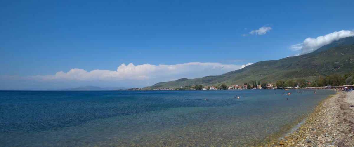 A clear blue sea with a rocky beach, a small town on the shore, and a mountain in the background.