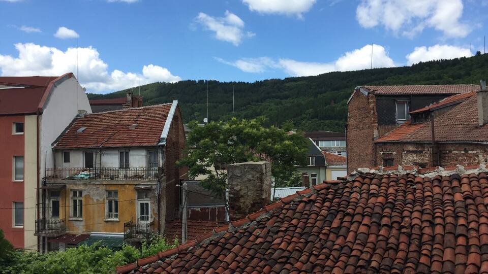 A hillside village with terracotta-tiled roofs, a mix of old and new buildings, and a clear blue sky with scattered clouds.