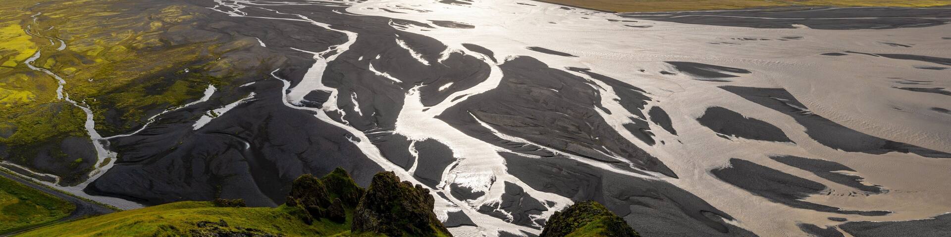A glacial landscape with a river flowing through it, surrounded by green fields and mountains under a cloudy sky.