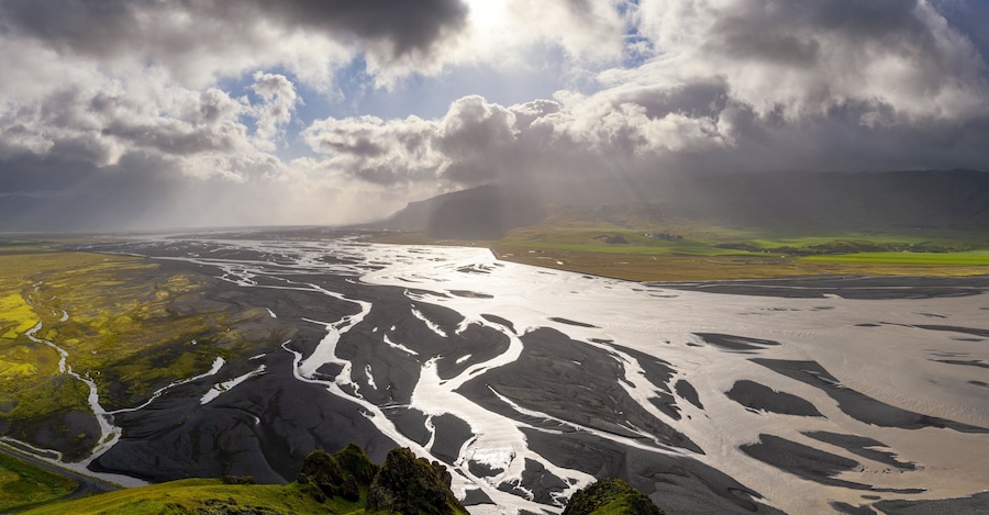 Un paisaje glaciar con un río que lo atraviesa, rodeado de campos verdes y montañas bajo un cielo nublado.