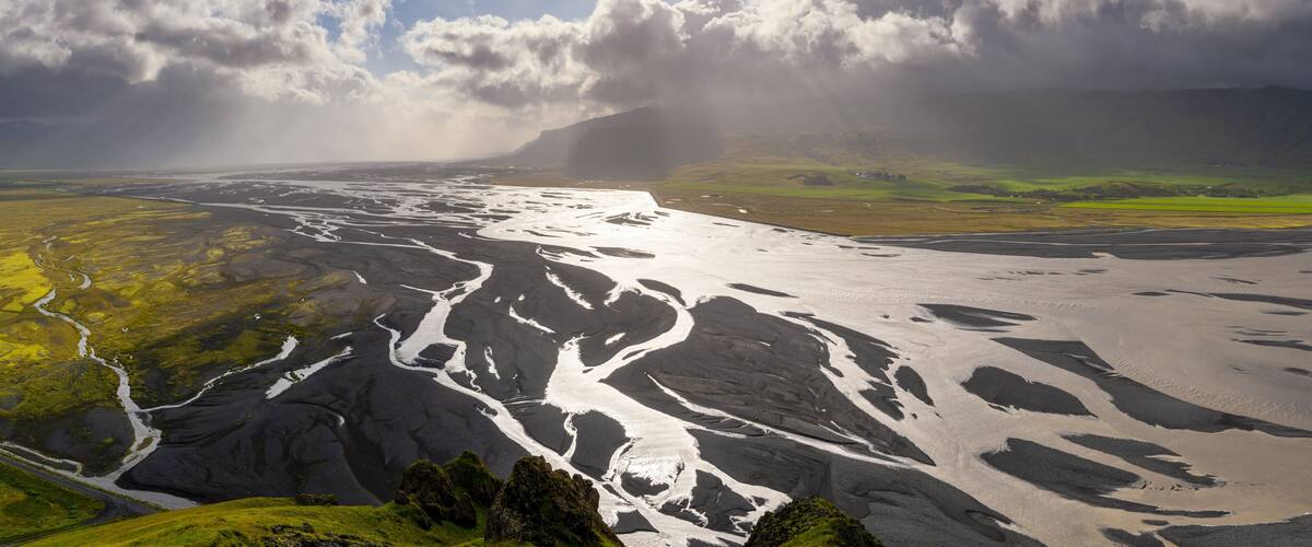 A glacial landscape with a river flowing through it, surrounded by green fields and mountains under a cloudy sky.