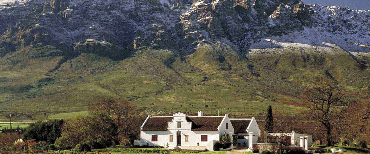 A white house with a stone wall and a vineyard in front, set against a snow-capped mountain.