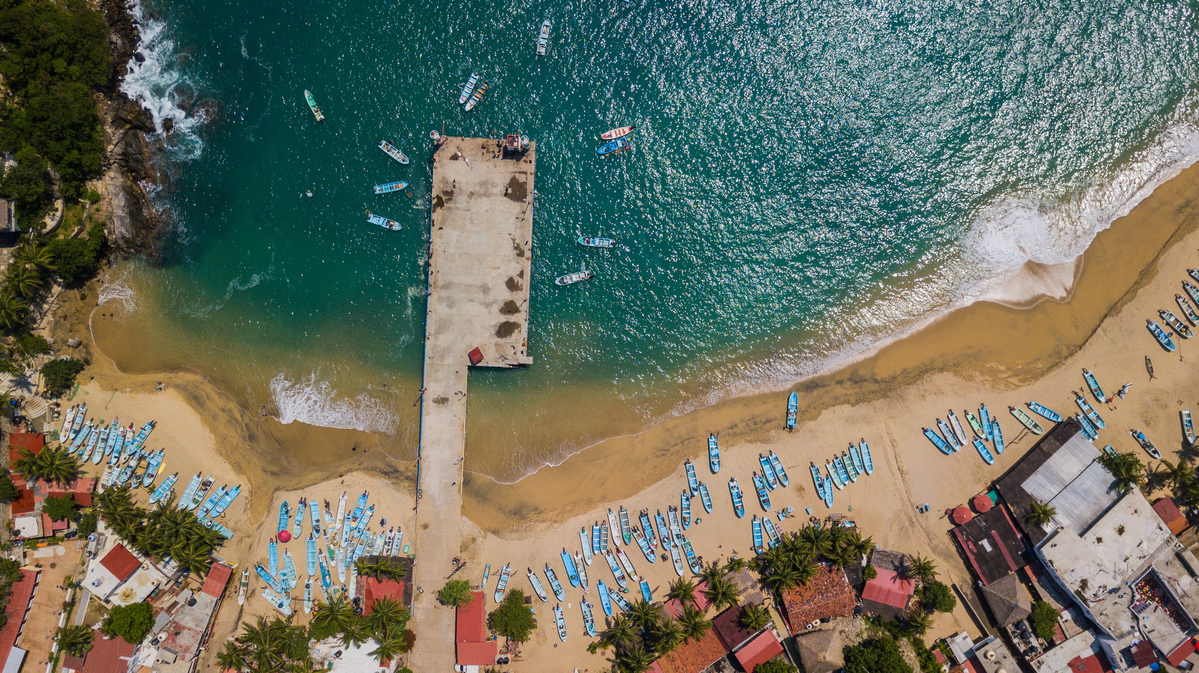 Vista aérea de una playa con numerosos barcos alineados a lo largo de la orilla y un muelle que se adentra en el agua.