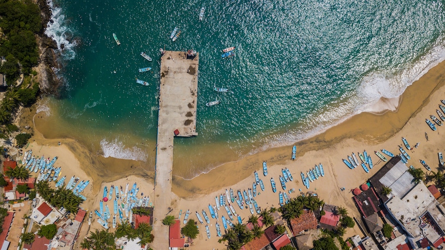 An aerial view of a beach with numerous boats lined up along the shore and a pier extending into the water.
