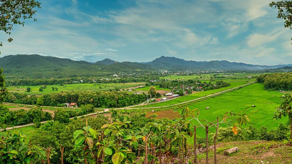 A lush green valley with terraced fields, a small village, and distant mountains under a partly cloudy sky.