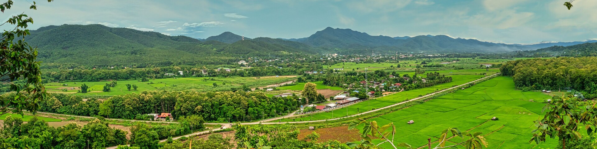 Un valle verde exuberante con campos en terrazas, un pequeño pueblo y montañas distantes bajo un cielo parcialmente nublado.