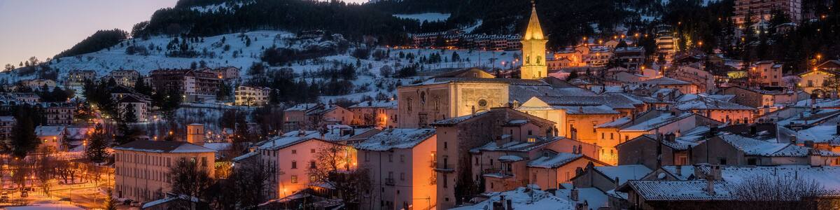 Une ville enneigée au crépuscule, avec des bâtiments illuminés et un clocher d’église imposant.
